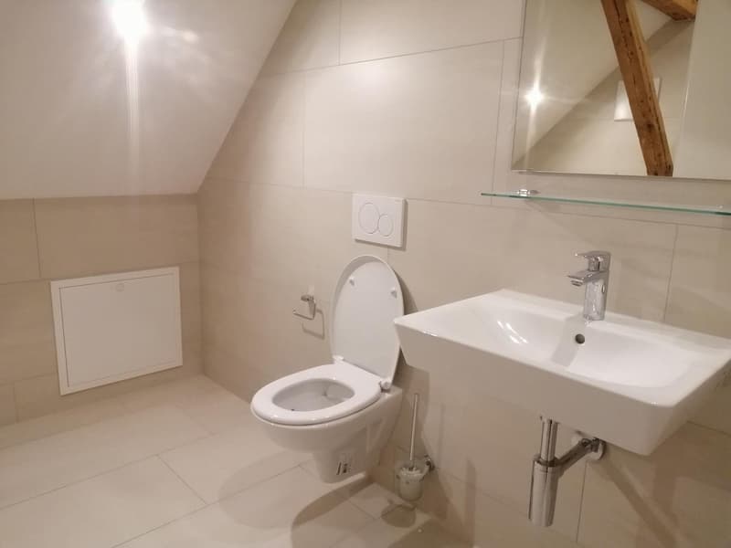 Modern bathroom with white tiled flooring, white sink, silver faucet, open toilet, mirror, and wooden ceiling