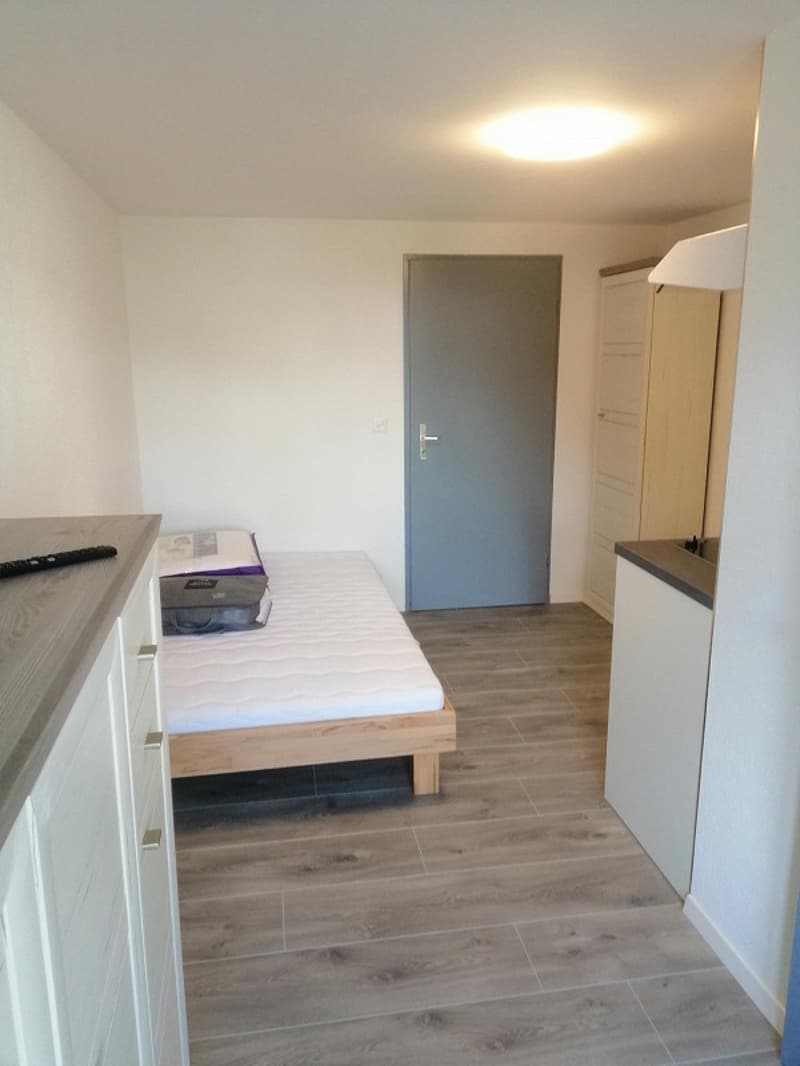 A small bedroom with a wooden bed, two white cabinets, tiled floor, and gray walls. There is an exit door in the center and a remote control on the cabinet.