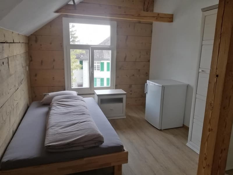 Small wooden bed, small bedside table, small white fridge, wooden walls, wooden floor