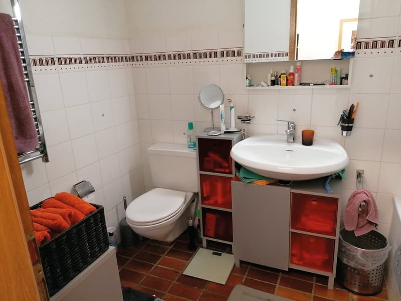 white tiled walls, white toilet, white sink, mirror, storage cabinet, towel rack, trash bin, towels