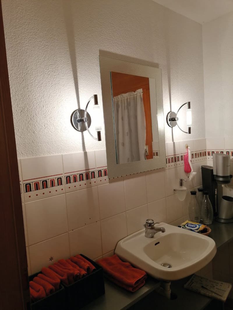 Bathroom with white sink, mirror, wall-mounted lights, tiled walls, towels, and a coffee machine.