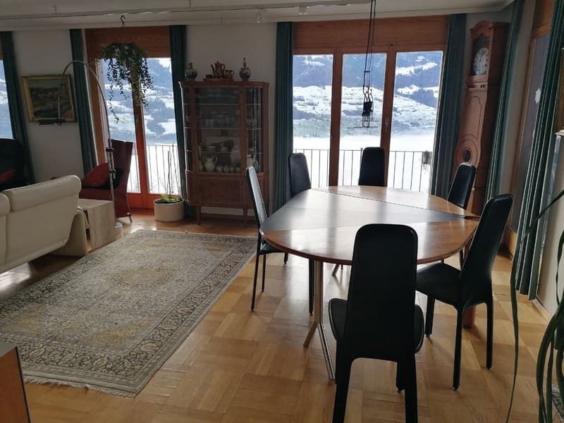 Dining area with a wooden table and chairs, wooden floor, large windows, balcony, rug, cabinet, and a clock