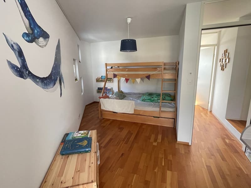 A room with wooden floor, white walls, bunk bed, whale wall art, wooden desk with books, and a lamp