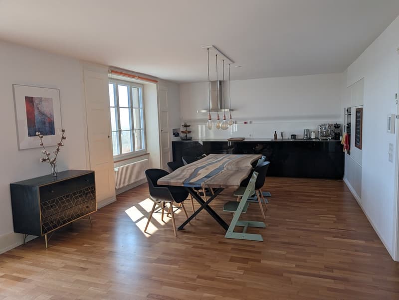 Dining room with wooden floor, large window, dining table, chairs, black cabinet, picture frame, radiator, kitchen with appliances