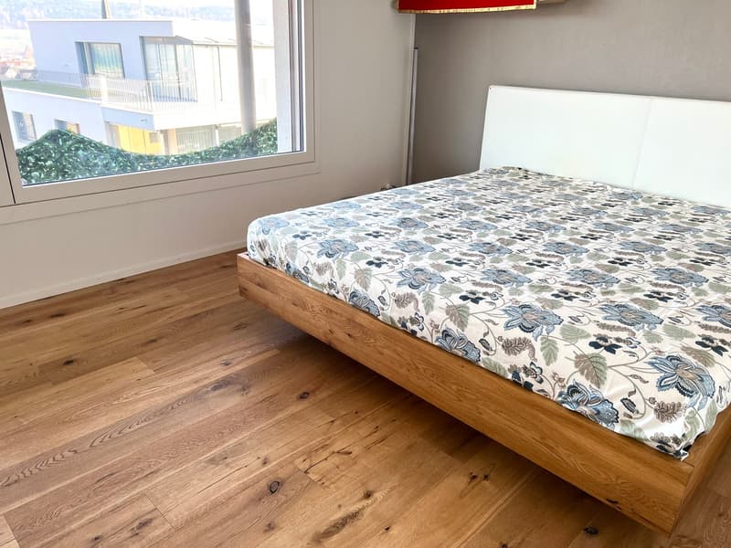 wooden floor, large window, bed with floral bedspread, white headboard