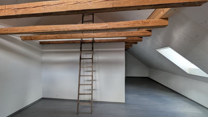 Empty attic room with wooden beams, a wooden ladder, and a white wall. The floor is tiled in gray.