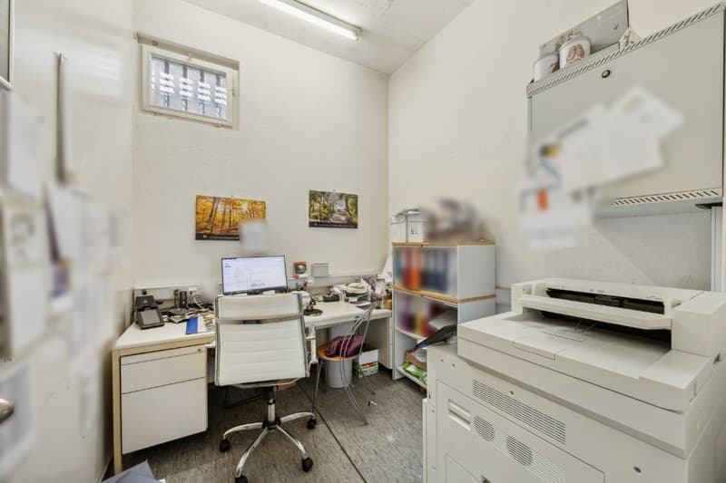 Small office space with a desk, computer, printer, and storage shelves. The room has white walls and a window, and appears to be used for administrative or work-related purposes.