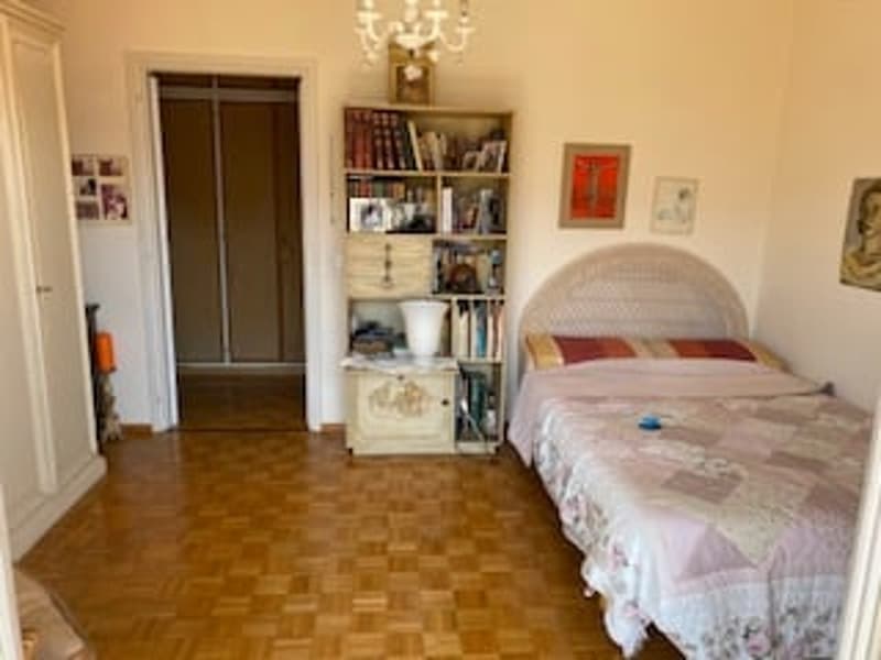 A bedroom with a bed, bedside table, and a bookcase filled with books and a lamp. The room has wooden floors and white walls.