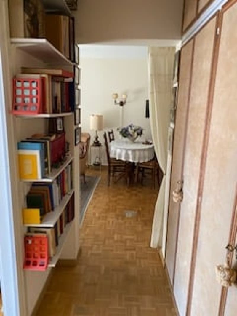 A hallway with wooden flooring, bookshelves filled with books, a dining table with chairs, and a lamp.