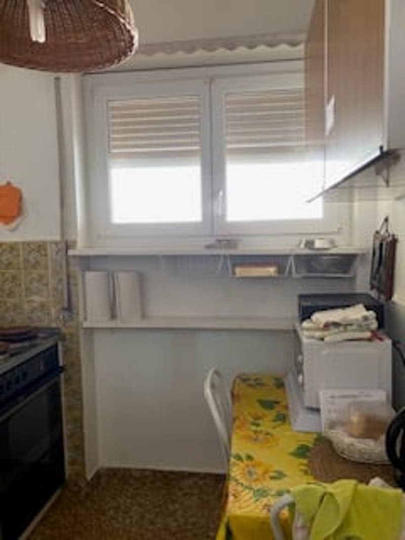 Small kitchen with white cabinets, a window, a stove, a microwave, a chair, a table, and a basket on the table.