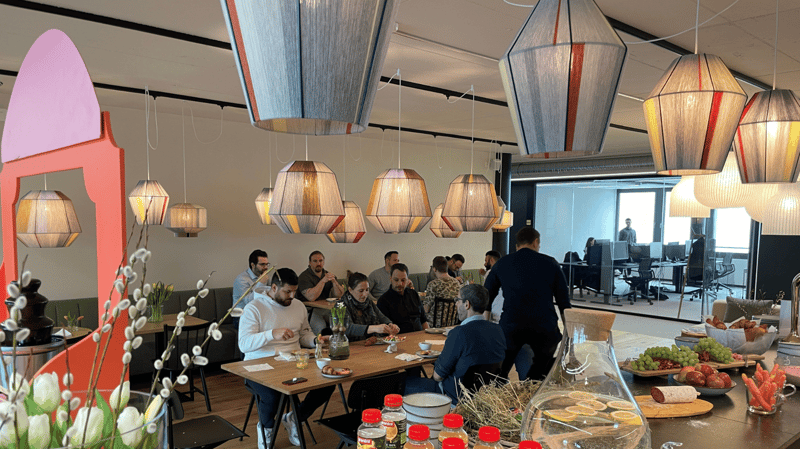 A group of people are sitting and standing in a room with wooden tables and chairs. The room has an open ceiling with hanging lamps. The lamps have a unique design. The room also has plants and flowers. The table is set with bowls of fruits, bread, and ot