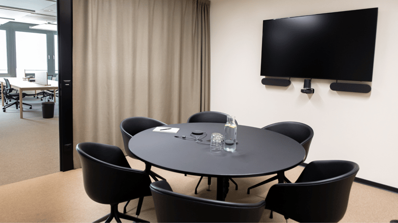 A small room with a round black table and black chairs around it, mounted TV, wooden table, curtains