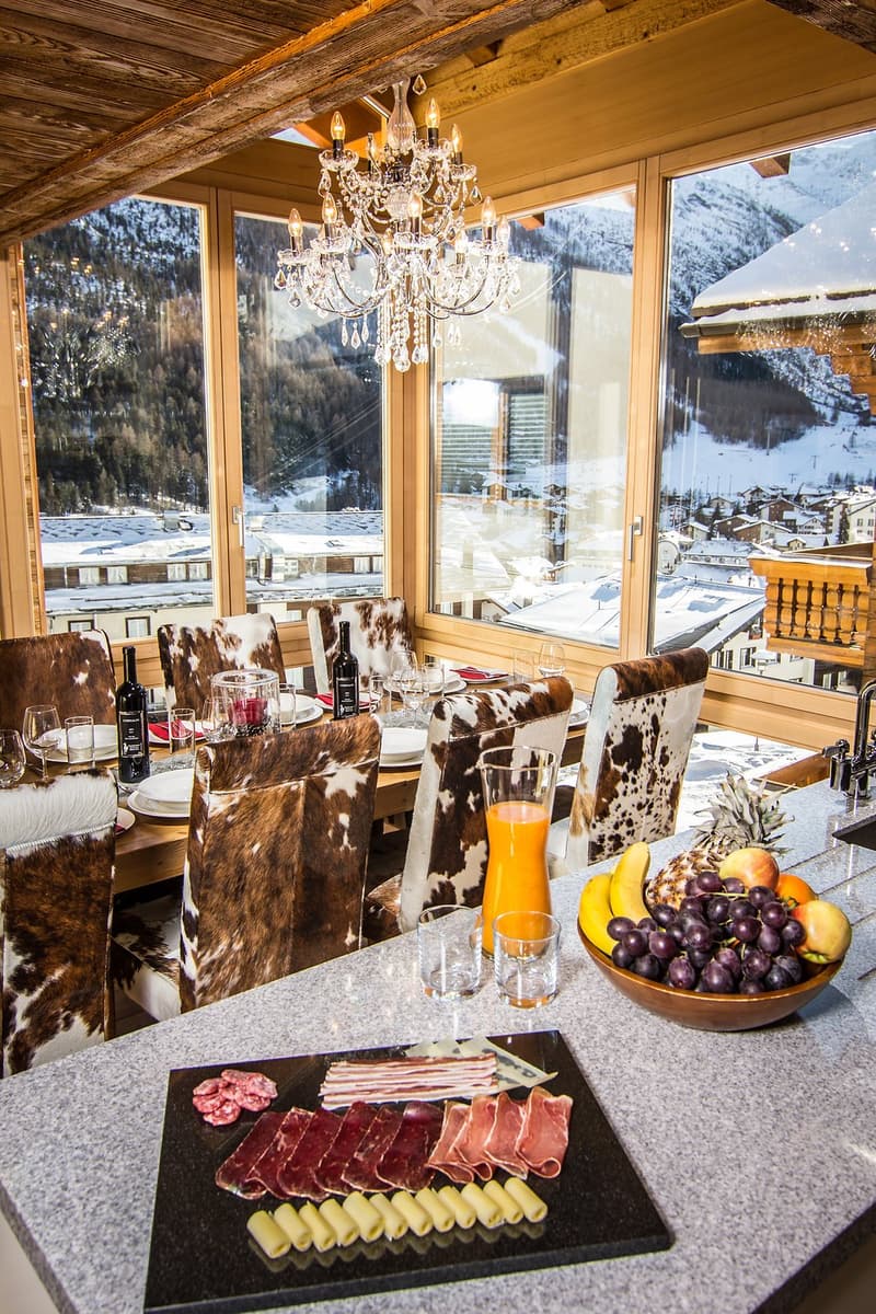 dining table set for six with bottles of wine, chandelier, glass wine glasses, a large bowl of fruit and a platter of charcuterie and cheese, view of snow covered mountains through large windows