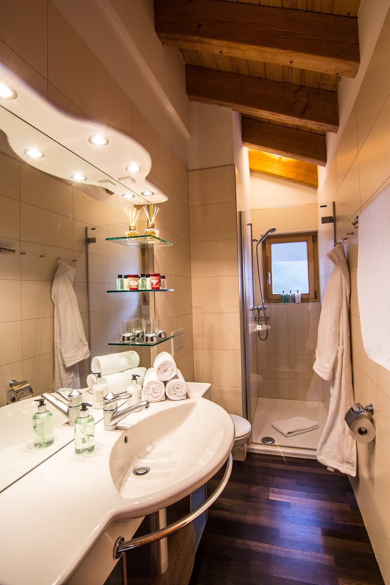 Contemporary bathroom with a white porcelain vanity, a shower stall, and decorative shelves with toiletries. The space also features wooden flooring and exposed wooden beams on the ceiling.