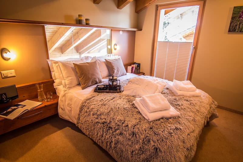 A large double bed with a fur cover, nightstand with a remote and books, and a lamp with wall sconces.