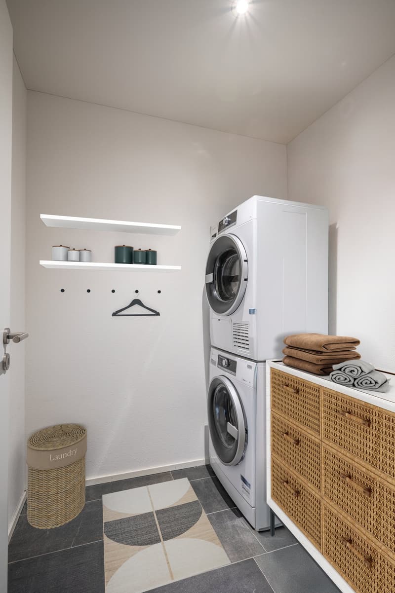 A small laundry room with a washing machine, a dryer, shelves, a wicker basket, and a rug.