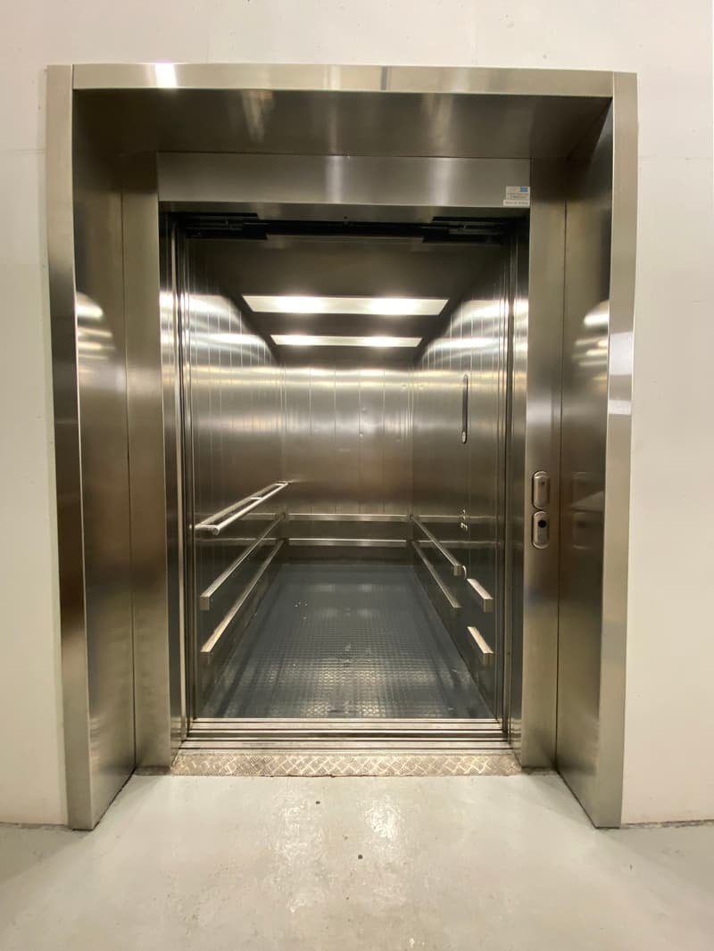 The image shows the interior of an elevator. The elevator has a stainless steel frame and doors, and the floor is made of a textured material. The elevator appears to be in a commercial or industrial setting.