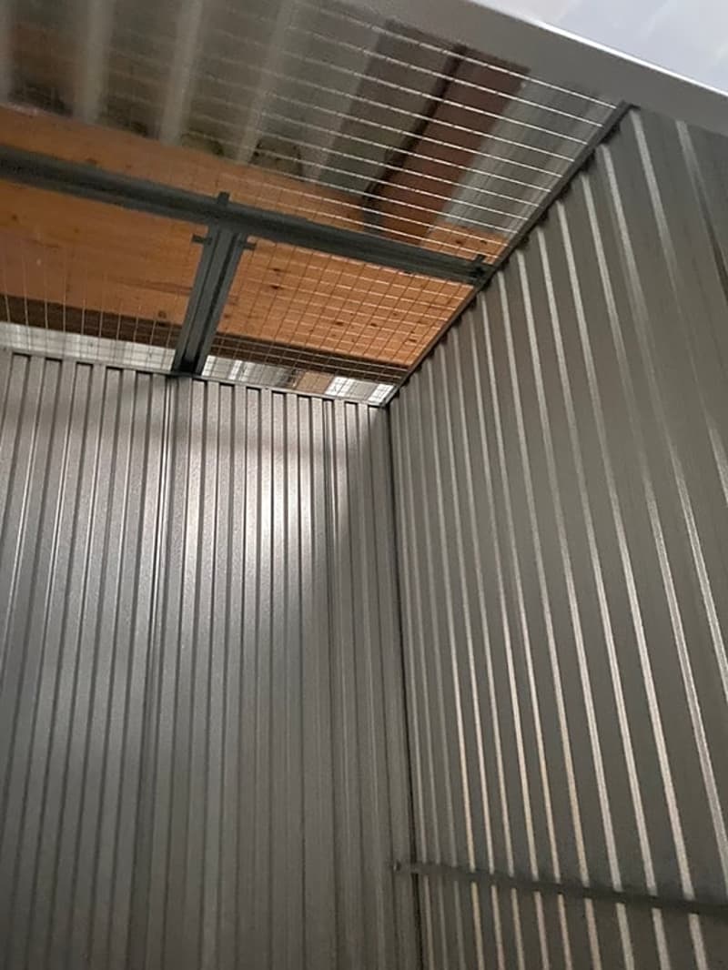 The image shows the interior of a garage or storage space. The walls are made of corrugated metal panels, and there is a wooden ceiling with metal support beams. The space appears to be designed for storage or housing a vehicle.