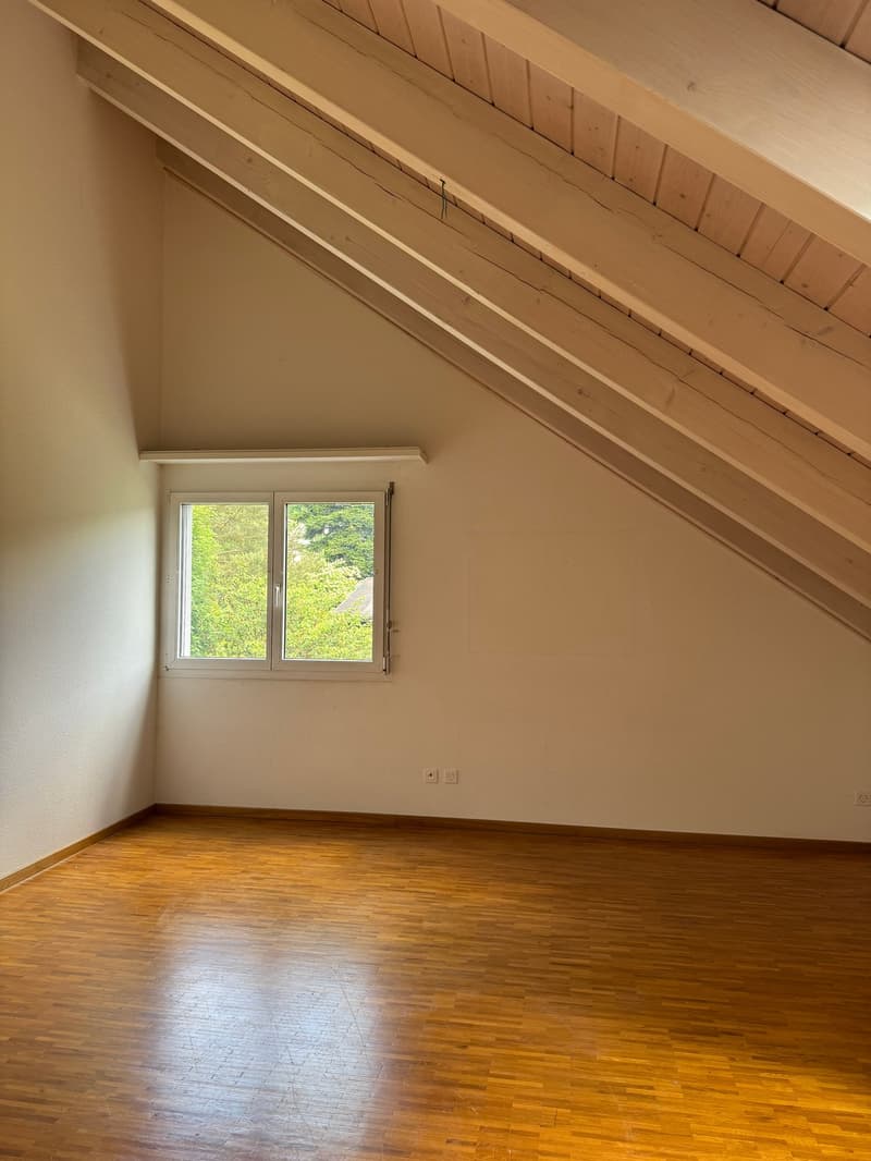 Spacious room with wooden floors, slanted ceiling with exposed beams, and a window overlooking the greenery outside.