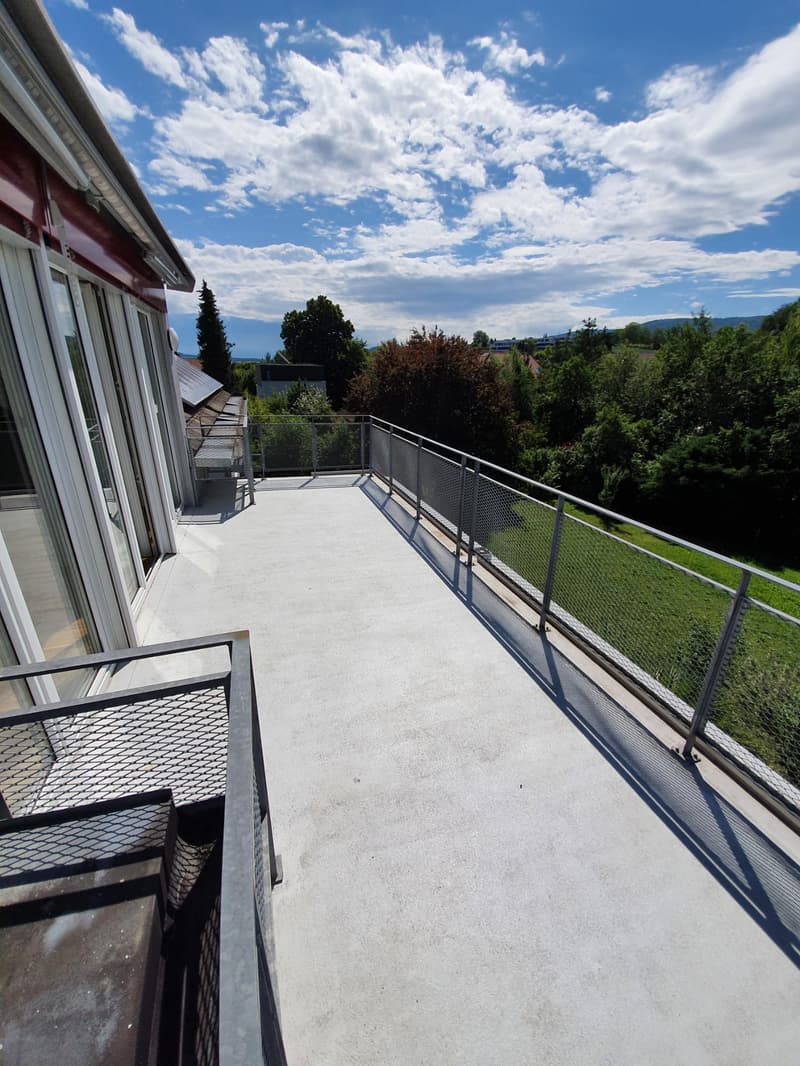 The image shows a spacious balcony or terrace with a concrete floor and metal railings. The balcony overlooks a scenic outdoor area with trees and greenery. The sky is filled with fluffy white clouds against a bright blue background.