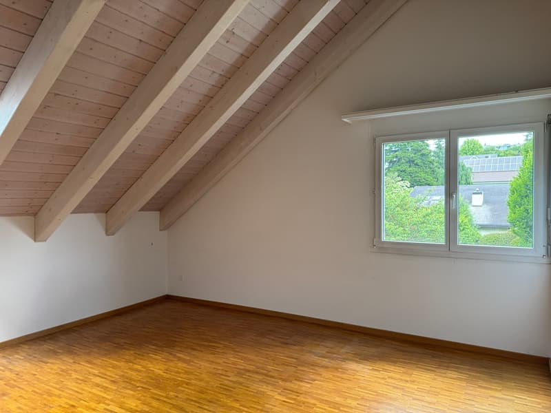 Attic room with wooden beamed ceiling, white walls, and hardwood flooring. Large window overlooking a green outdoor area.
