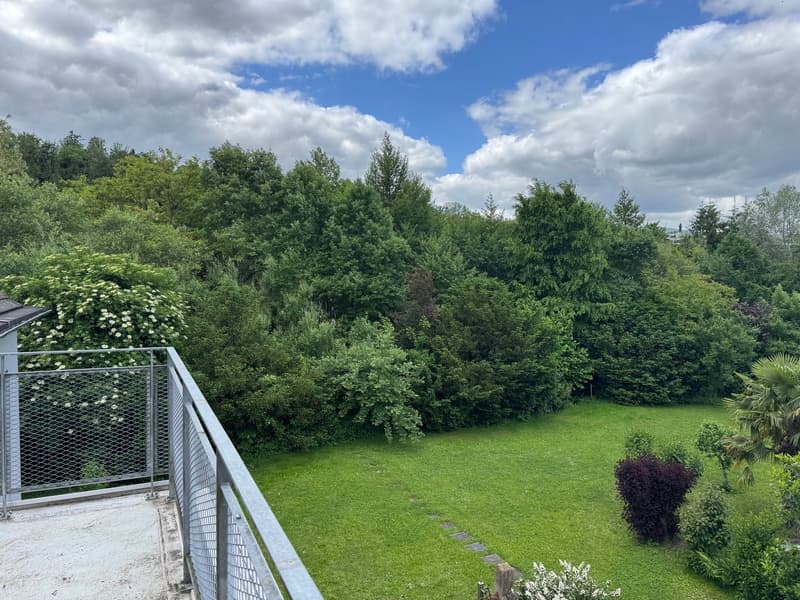 The image shows a lush, green garden with a variety of trees and shrubs. There is a balcony or terrace visible in the foreground, overlooking the expansive lawn and landscaping. The sky is partly cloudy, creating a picturesque outdoor scene.
