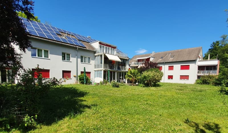 A multi-story apartment building with solar panels installed on the roof. The building has a green lawn and landscaping in the foreground, with some red accents on the windows and balconies.