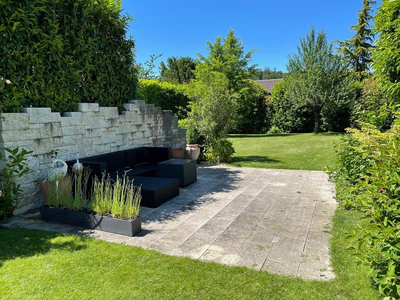 Outdoor patio with a stone wall, black lounge furniture, planters, lush green grass, and a scenic view.