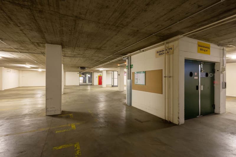 Large indoor parking garage with concrete floors and ceiling, yellow markings on the floor, and storage rooms or utility spaces along the walls.