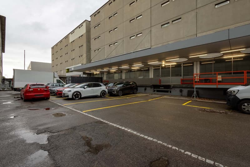 Large commercial building with a parking lot in front. The building has a gray exterior with multiple floors and windows. There are several cars parked in the lot, including a red car, a white car, and a black SUV.