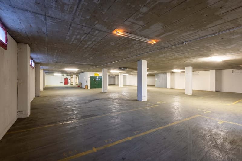 Large open space with concrete floor, white walls, and exposed ceiling with lighting fixtures. The space appears to be a parking garage or similar utility area with designated parking spots marked on the floor.