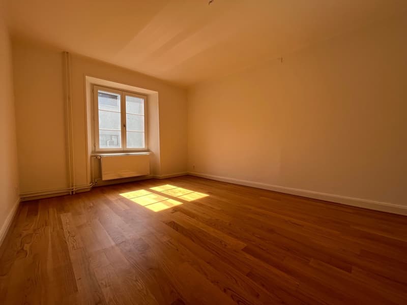 Empty room with wooden floor, white walls, large window, and a radiator.