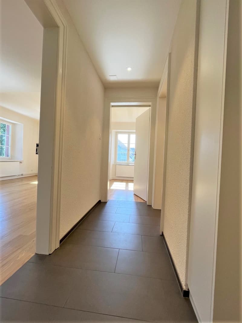 An empty hallway in a home with wooden floors and white walls. There are two doors on both sides leading to other rooms. The floor is tiled and shiny.