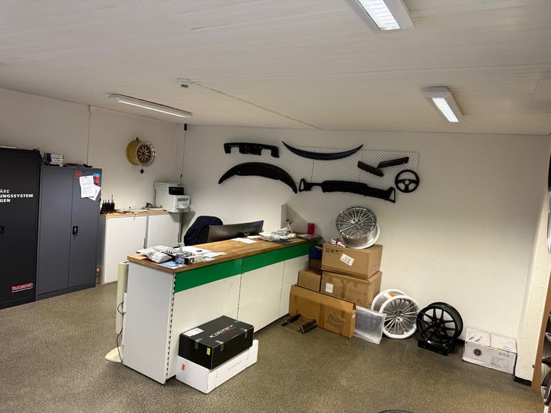 An office space with a desk, chair, cabinets, storage boxes, black items on the wall, monitors, lamps, tires, and wheels.