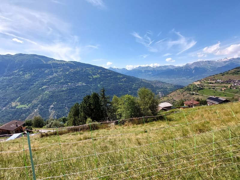 The image shows a scenic mountain landscape with rolling hills, lush green vegetation, and a small village nestled in the valley below. The sky is clear and blue, with wispy clouds. The mountains in the distance appear snow-capped. There is a fence or wir