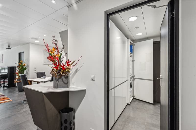 Modern office space with white walls, recessed lighting, and a decorative floral arrangement on a marble-topped reception desk. The room features a glass-paneled door leading to an adjacent area.