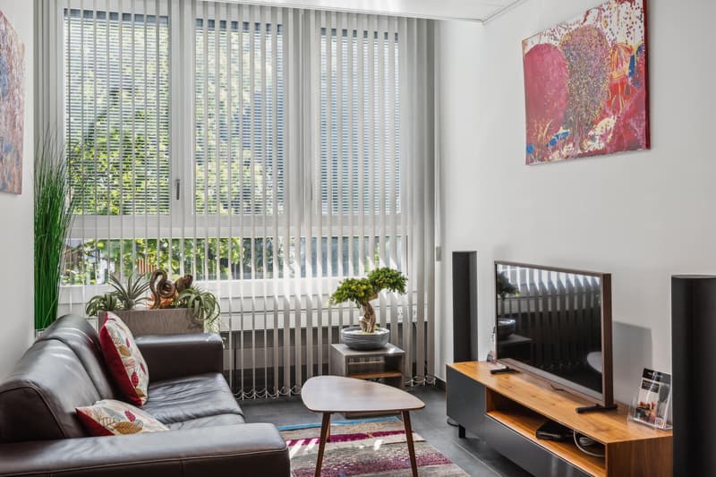 Spacious living room with large windows, vertical blinds, leather sofa, coffee table, TV stand, and colorful artwork on the walls. The room has access to a balcony or terrace, providing natural light and a view of the outdoors.