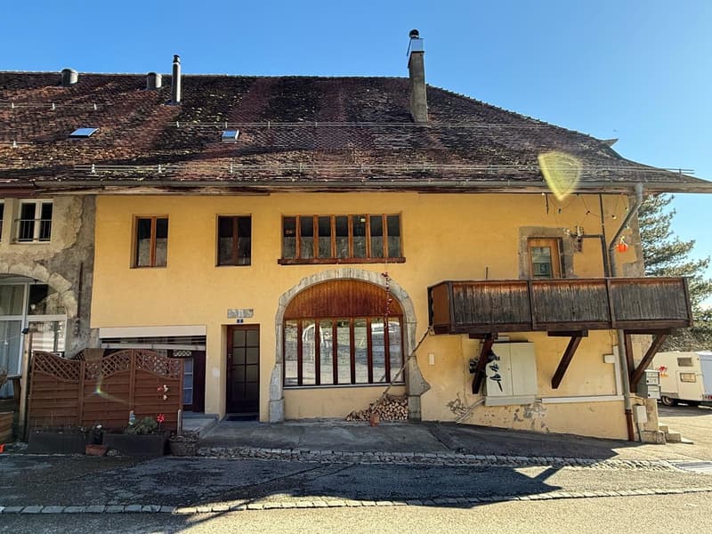 Stone and yellow walls, two storey, tiled roof, wooden balcony, arched doorway, chimneys, windows with metal bars, garden gate, several trees, stack of firewood, road with pebbles