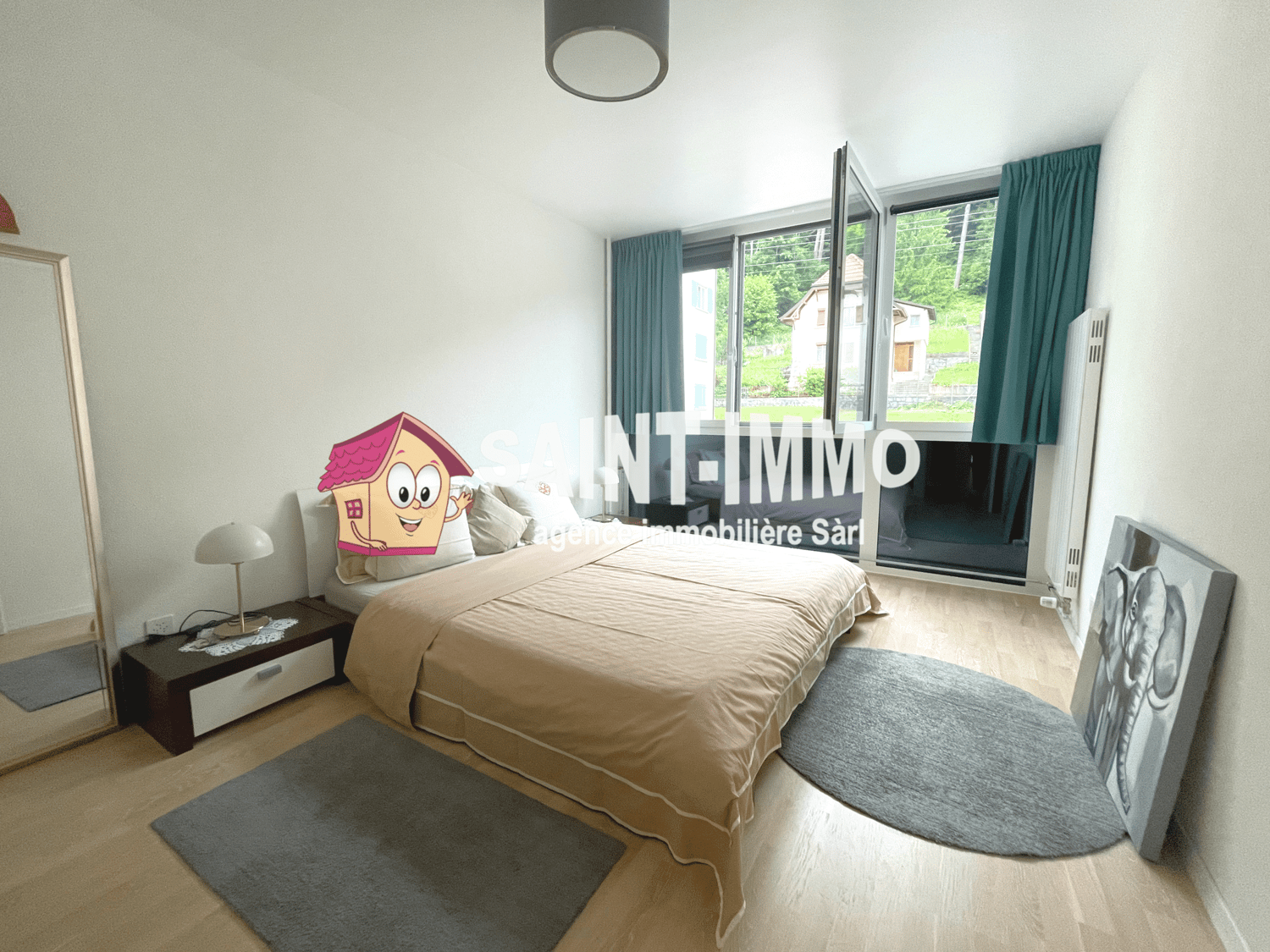 Spacious bedroom with large windows, hardwood flooring, beige bedding, and a decorative pillow in the shape of a house. The room also has curtains and a rug.