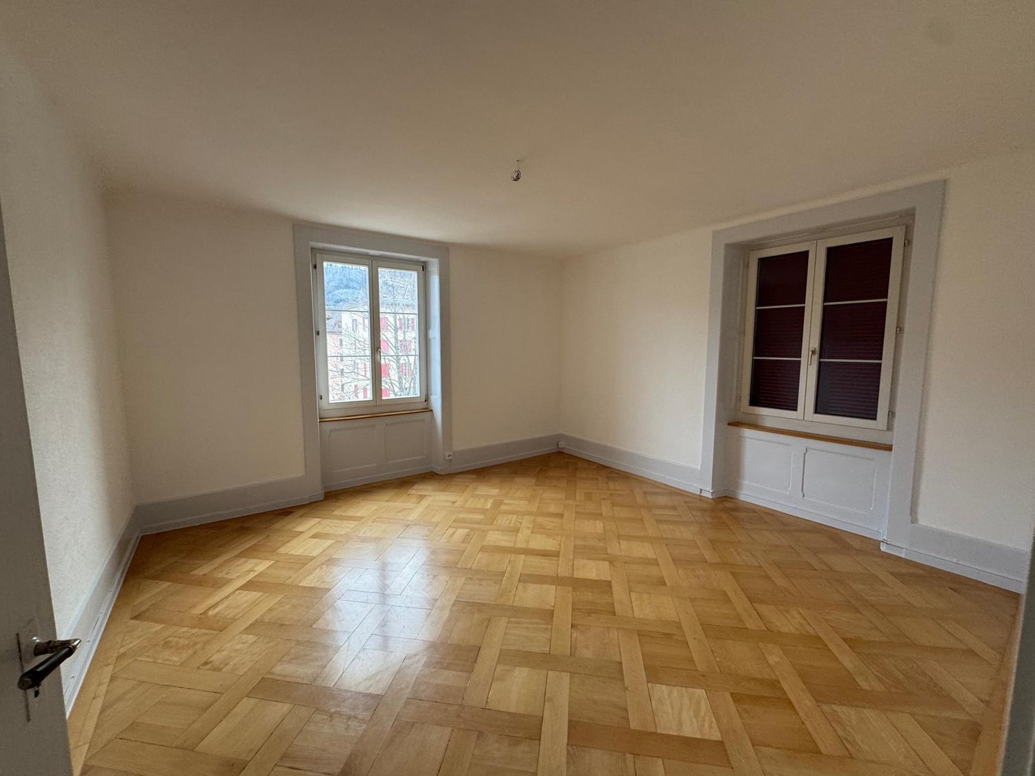 This image shows a spacious room with hardwood parquet flooring and white walls. The room has a large window that provides natural light, and there is a built-in cabinet or shelving unit along one of the walls.