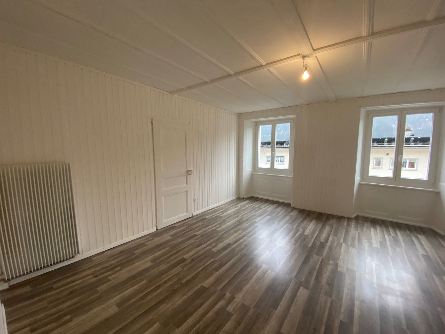 Empty room with wooden floor, white walls, 2 windows, door, radiator