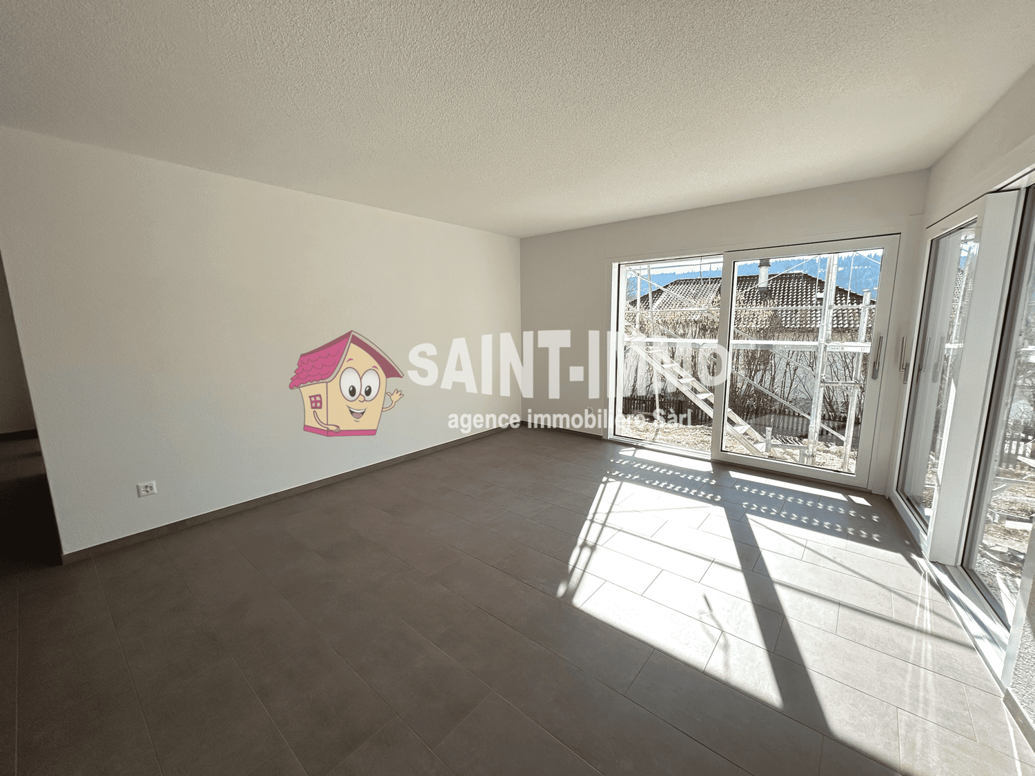 Large open room with gray tile flooring, white walls, and large windows providing natural light. The room features a prominent Saint-Immo real estate agency logo on the wall.