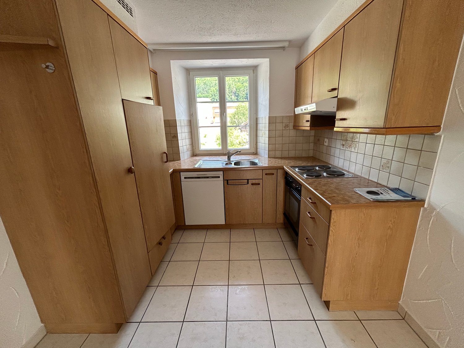 Galley-style kitchen with wooden cabinets, tile flooring, stove, sink, and dishwasher. Large window providing natural light.