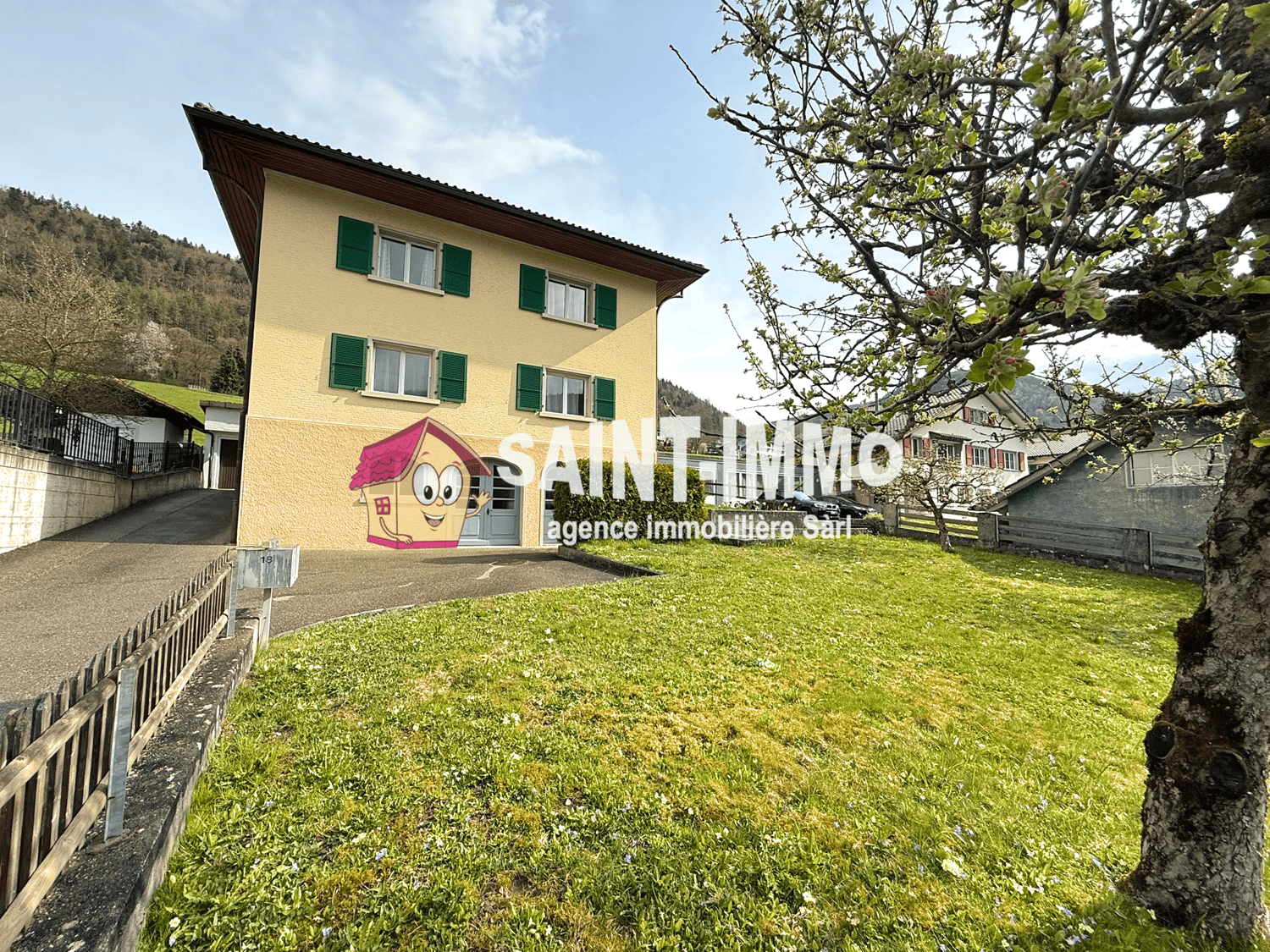 Yellow house with green shutters, driveway, grassy lawn, tree, mountains in the background