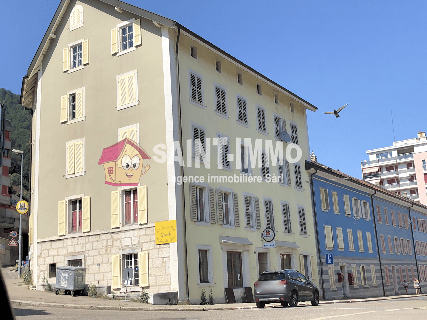 A multi-story building with a cartoon character on the side, yellow shutters, and a sign for an agency. A car is parked in front.
