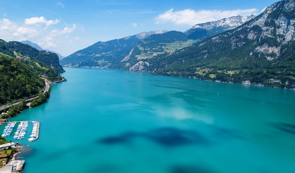 Aerial view of a scenic lake surrounded by mountains, with a marina and boats visible on the shore. The lake has a vibrant turquoise color and the landscape is lush and green.