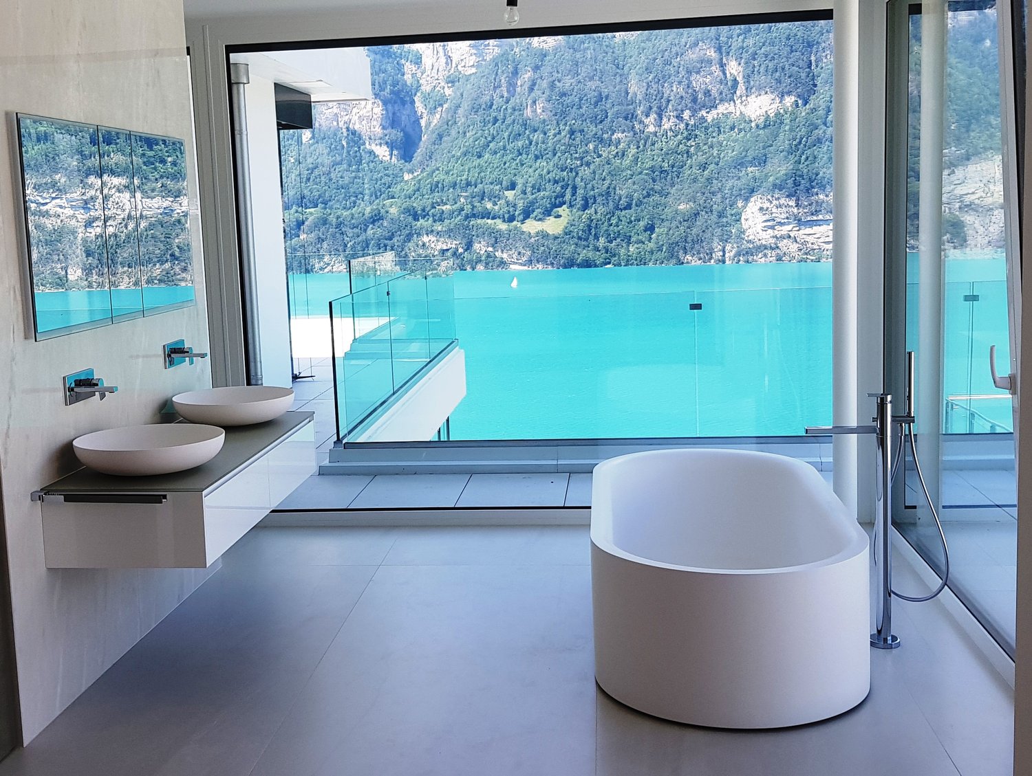 Spacious bathroom with large windows offering a stunning view of the turquoise lake and surrounding mountains. The bathroom features a modern design with a freestanding bathtub, double vanity sinks, and a glass-enclosed shower area.