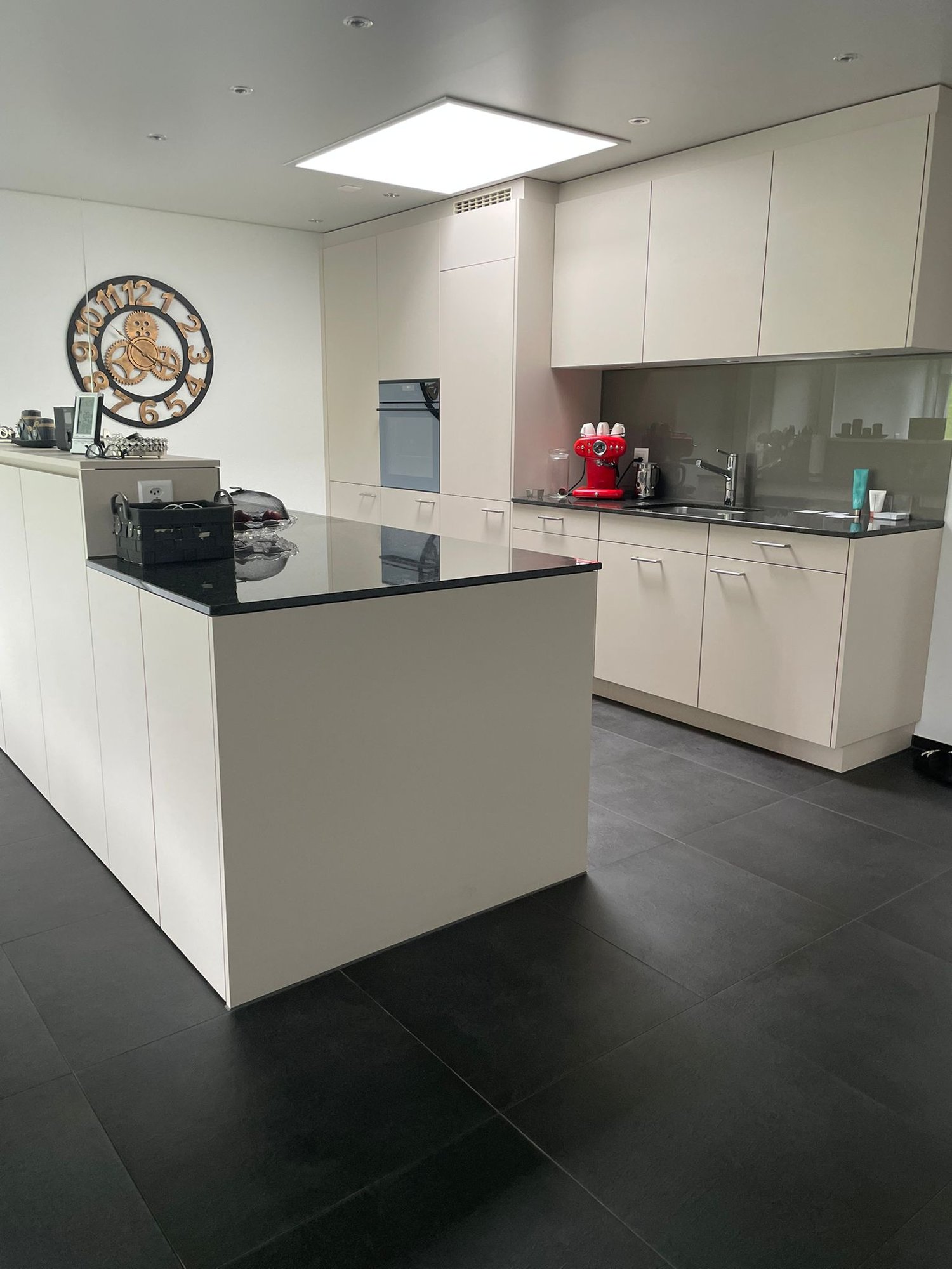 modern kitchen with white cabinets, black countertops, built-in oven, large island, sink, faucet, coffee machine, countertop storage, clock, large clock hanging on wall