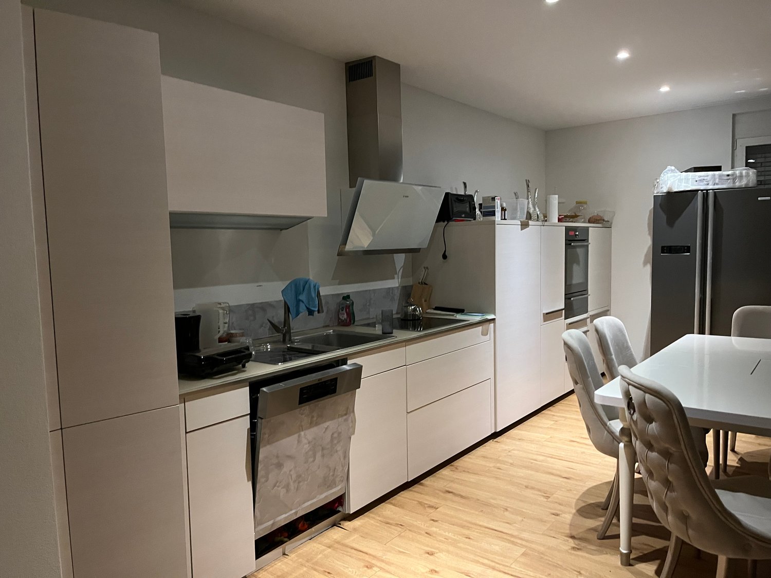 white modern kitchen with built-in oven, dishwasher, refrigerator, kitchen island, wooden floor, breakfast table with chairs