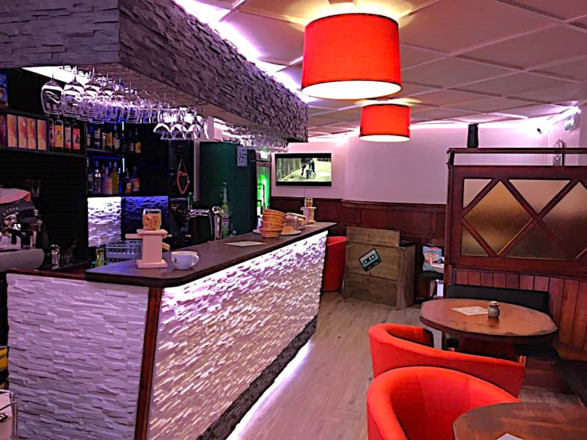 The image shows the interior of a bar or restaurant. The space features a stone accent wall, a long bar counter with shelves displaying various liquor bottles, red pendant lights, and round wooden tables with red chairs. The space appears to have a cozy a